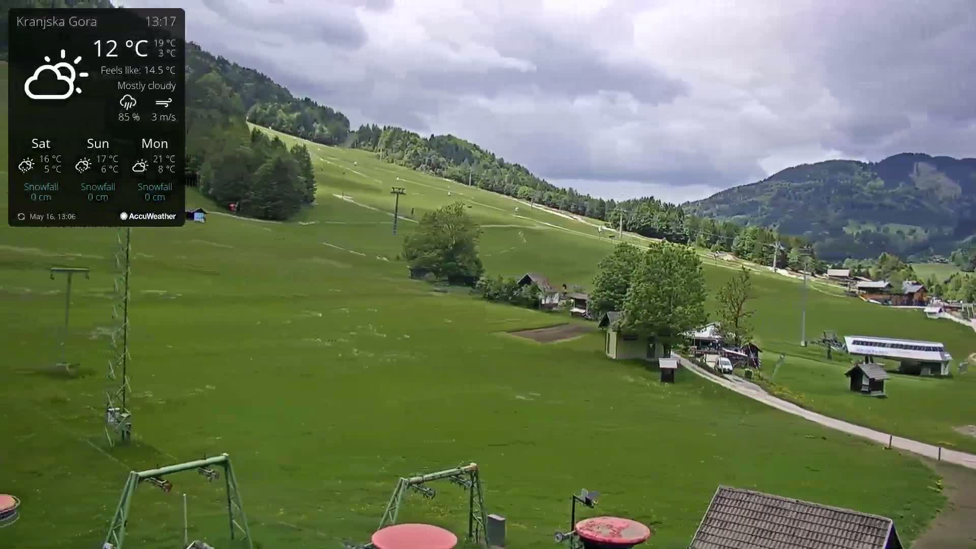 A mostly cloudy day reveals a grassy hillside with ski lifts and a few buildings, nestled against a backdrop of mountains.