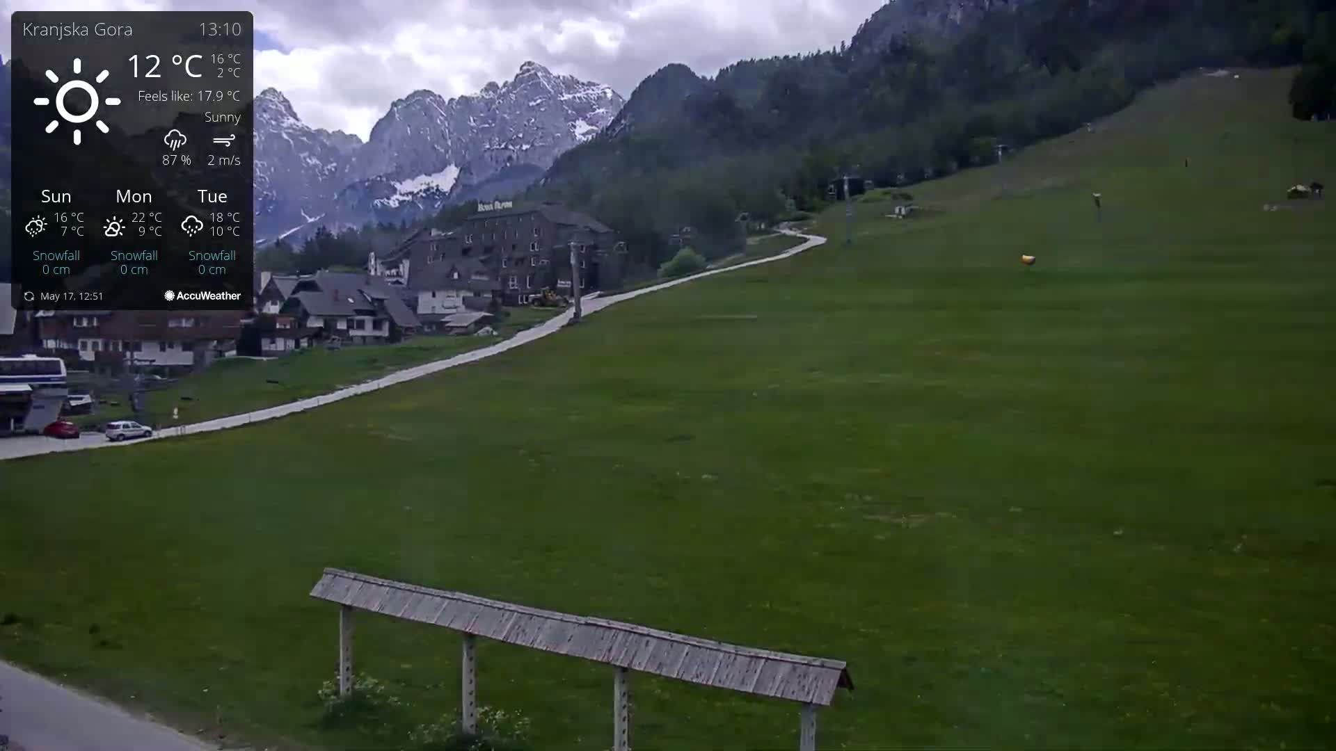 A sunny mountain landscape with green grassy slopes, a road, a few buildings, and a chairlift in the background under a partly cloudy sky.