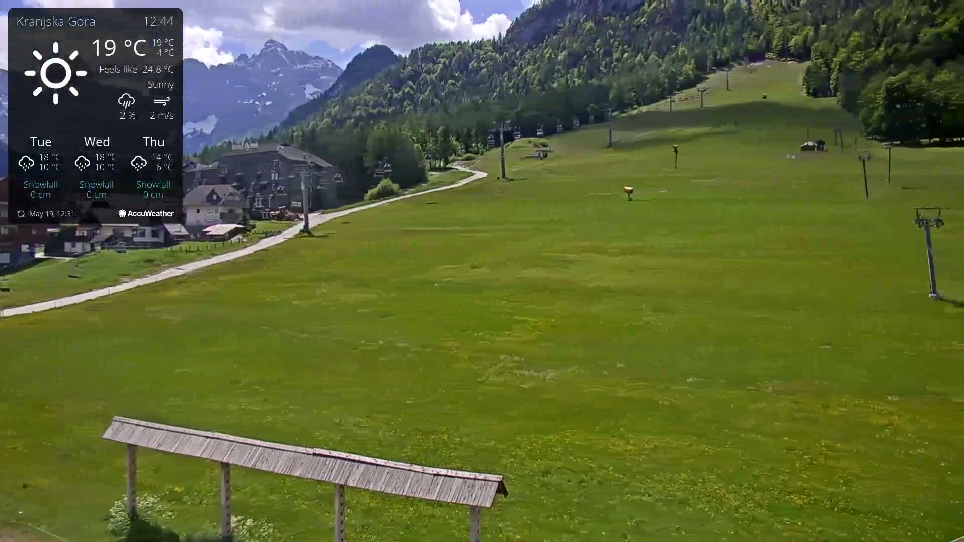 A sunny, grassy mountain slope with a few buildings and ski lift infrastructure is visible under a mostly clear sky.