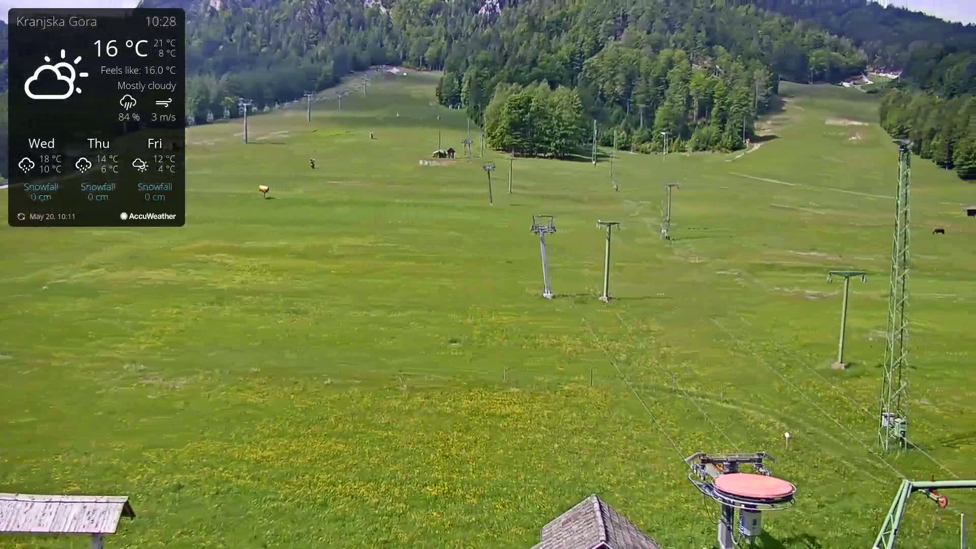 A mostly cloudy,  daytime view of a grassy field with ski lift infrastructure, and a forested hillside in the background.