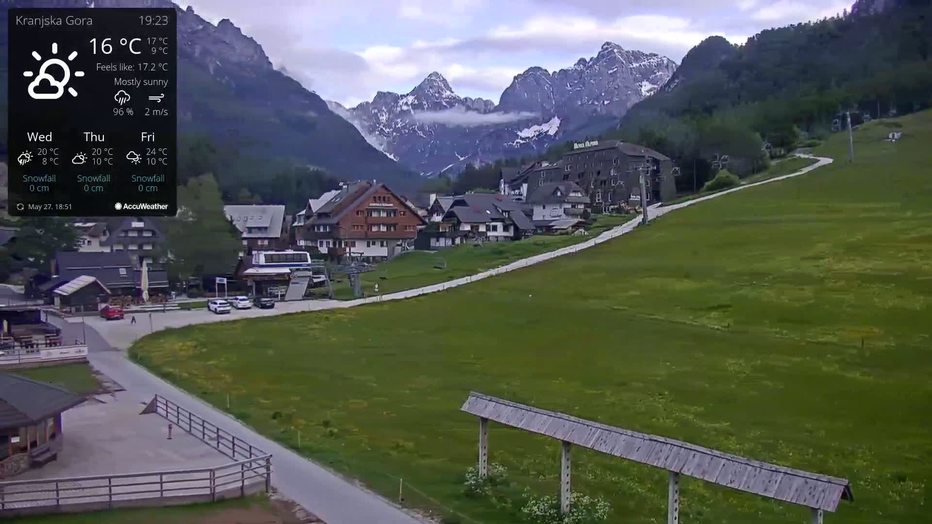 A mostly sunny mountain village scene shows several buildings, a road, and a ski lift, with mountains in the background.