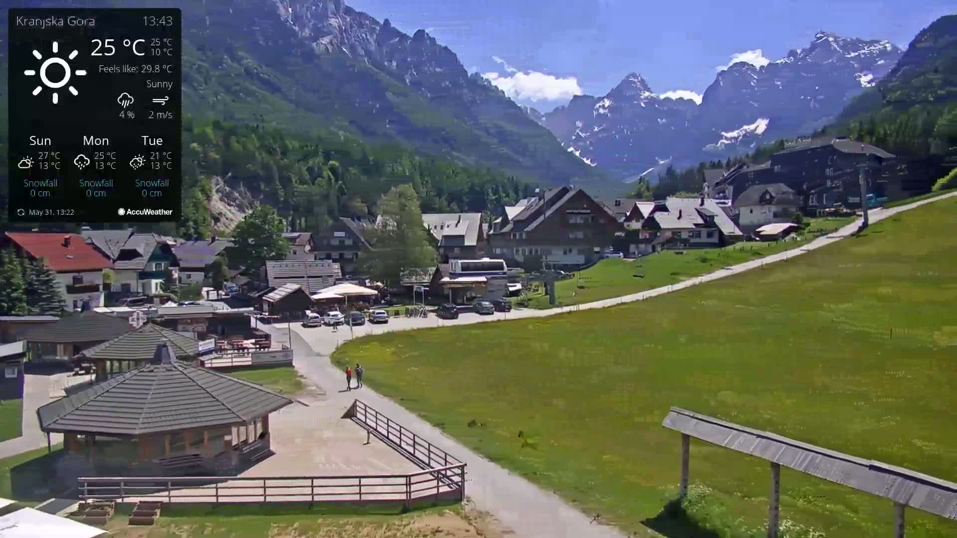 A sunny mountain village with a paved road winding through green fields, nestled against a backdrop of towering, snow-capped mountains.