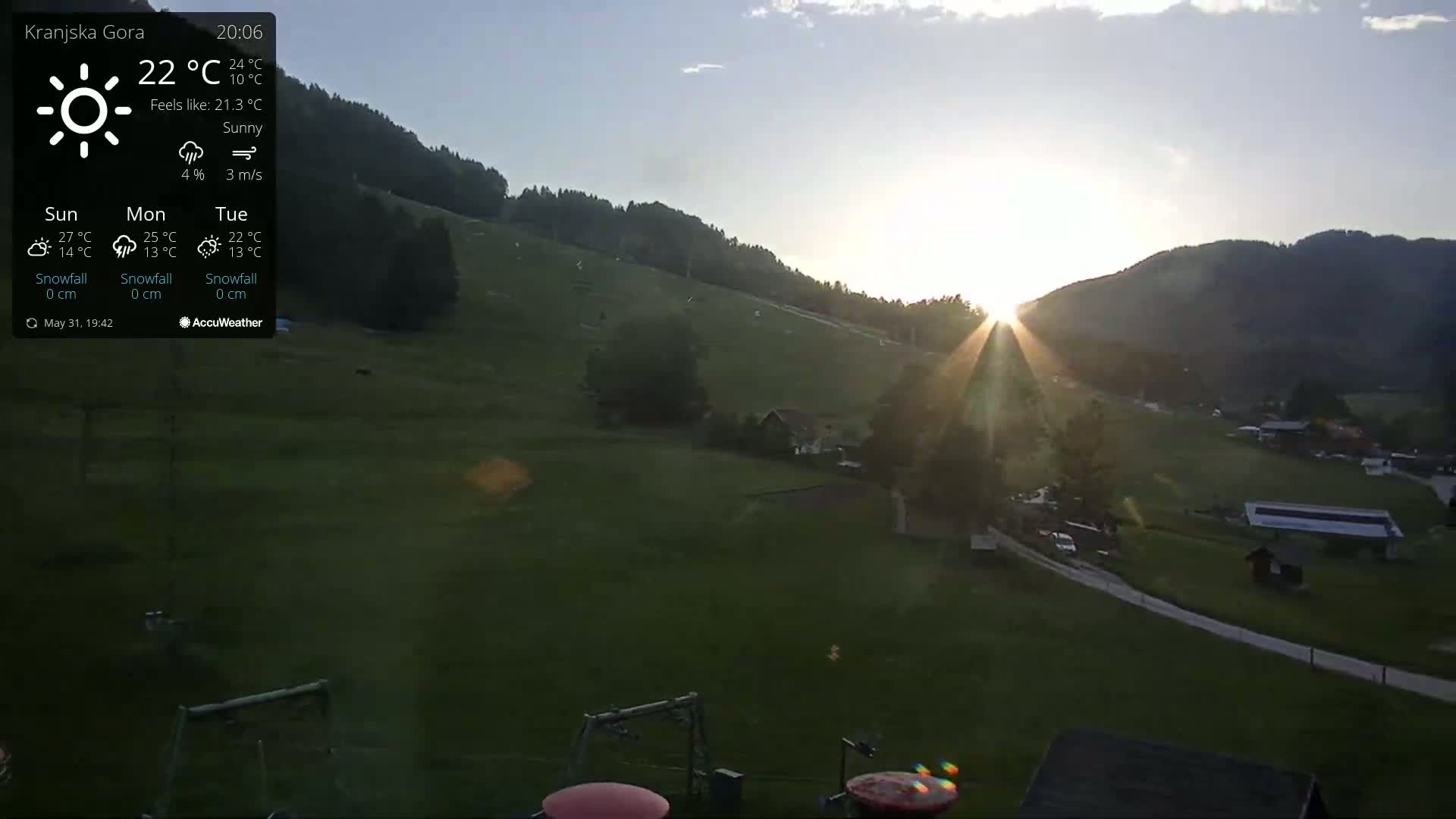 A sunny sunset casts rays over a green grassy hillside with buildings and ski lifts visible in the foreground.