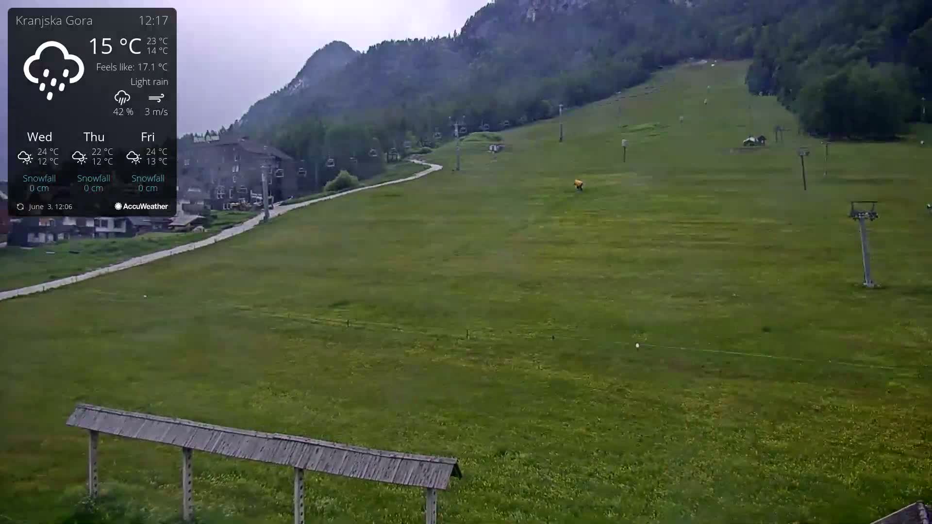 A grassy hillside with ski lifts and a building in the background under light rain.