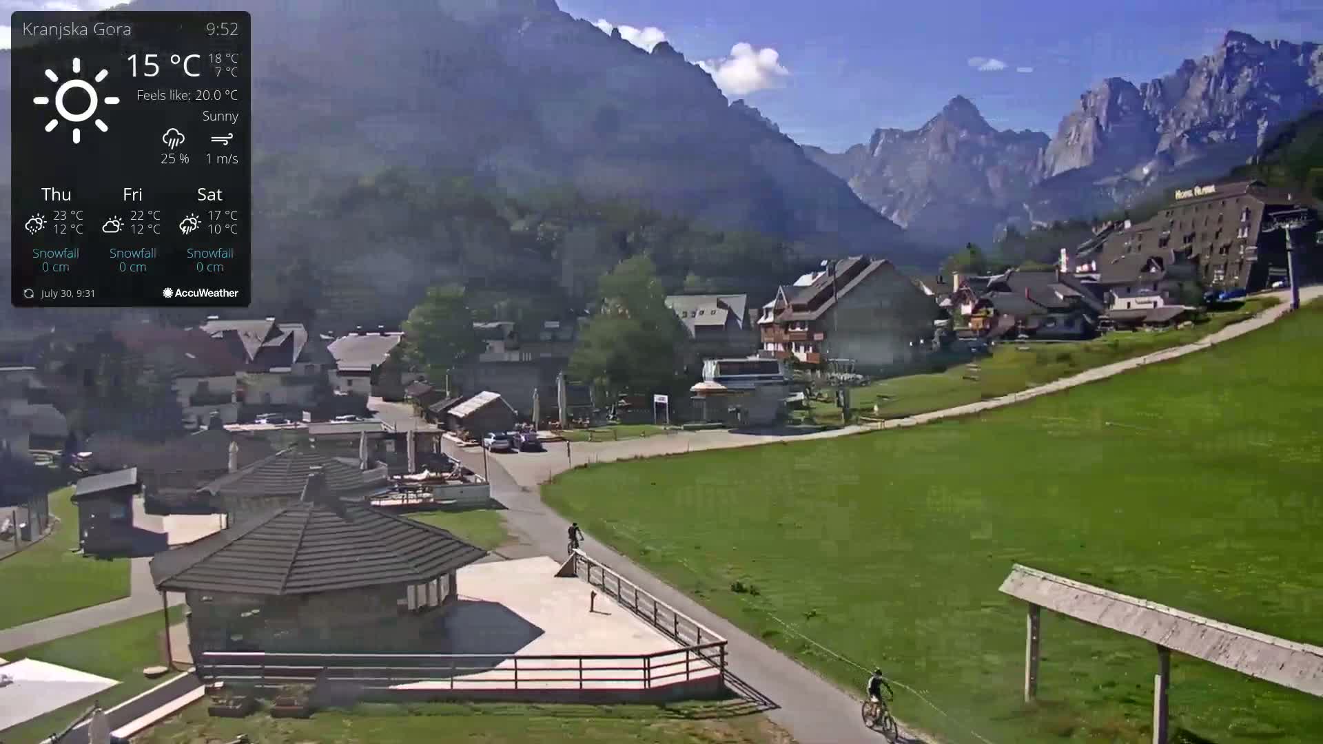 A sunny mountain village scene with two cyclists on a path, showing a few buildings and a grassy area beneath towering mountains.