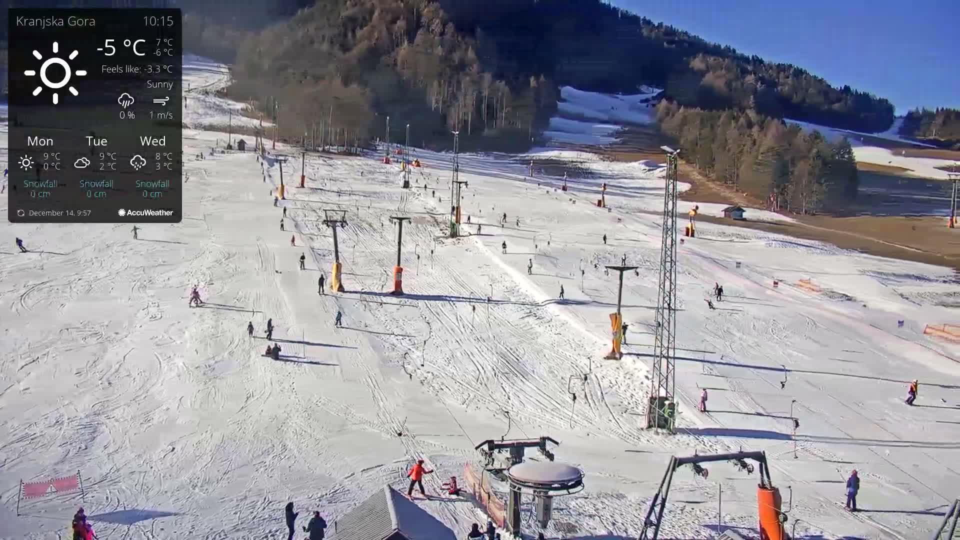 On a cold day with light snow, a snow-covered ski slope is visible, featuring numerous ski lifts, a bordering winter forest, and a few people along with a snow cannon.