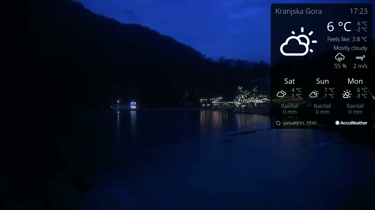 A nighttime view of a mostly cloudy scene shows a dark body of water reflecting the lights of a structure on the far shore, with dark mountains rising behind it.