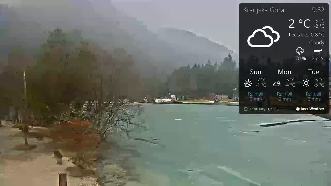 A partly frozen lake is seen from a lakeside vantage point, under a cloudy sky with misty mountains in the background.