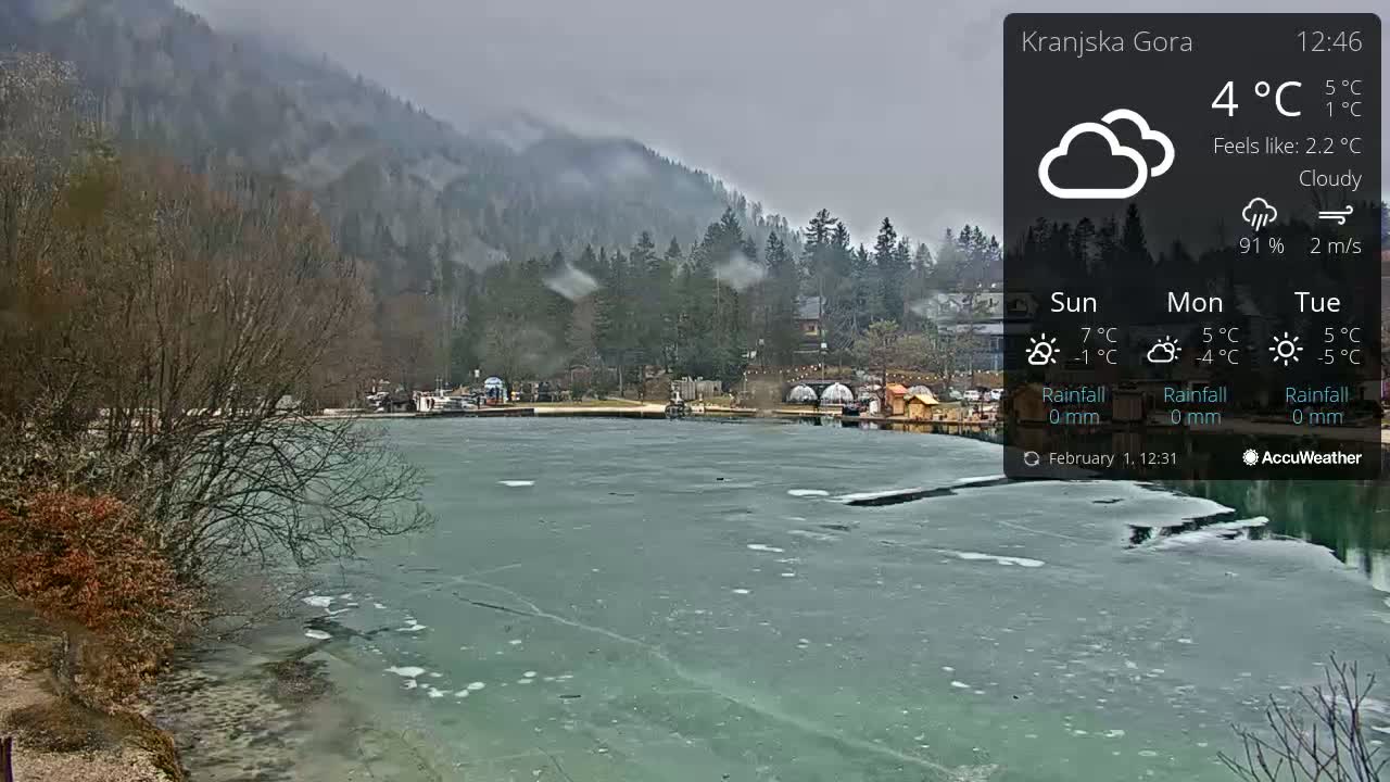 A partially frozen lake is seen under a cloudy sky, with a mountain range in the background.