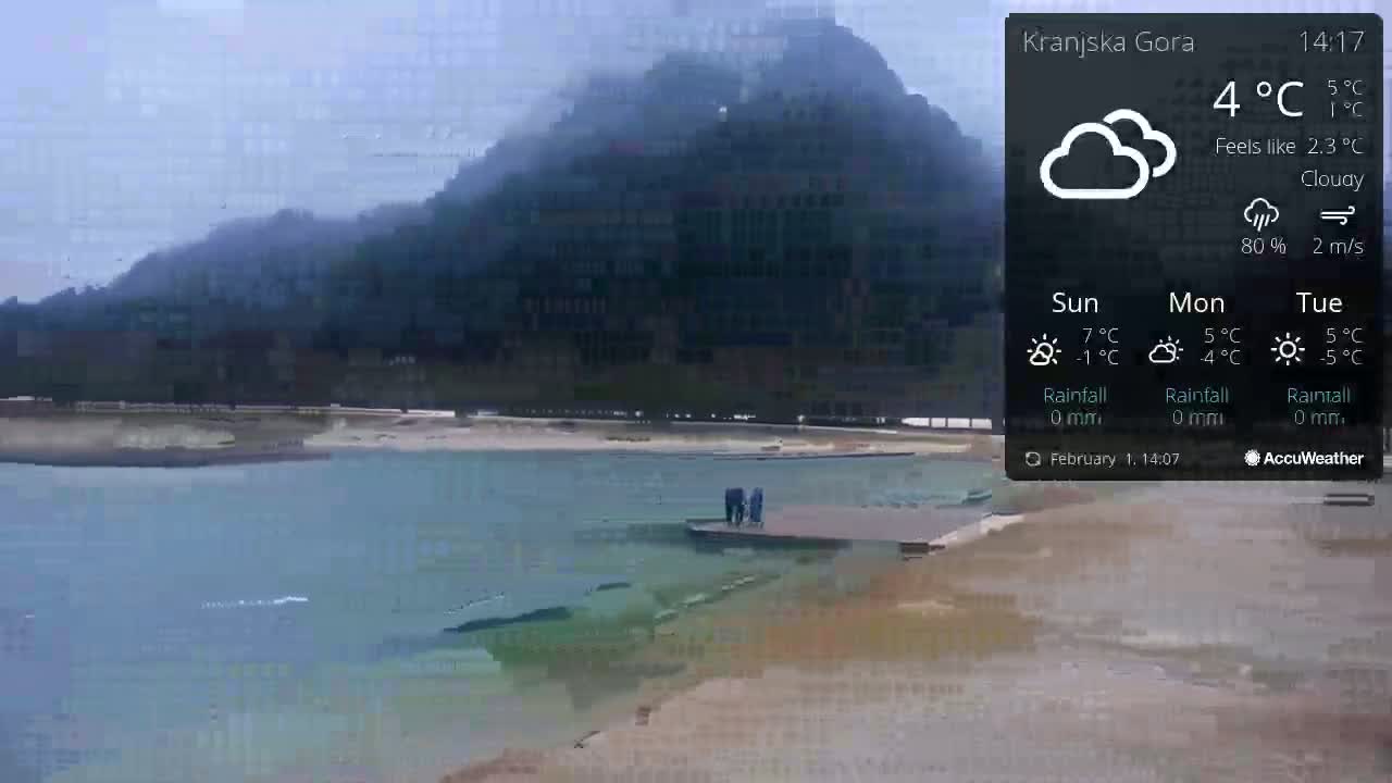 A cloudy and calm day shows a body of water next to a sandy shore with a mountain in the background, and a few people standing on a platform in the water.