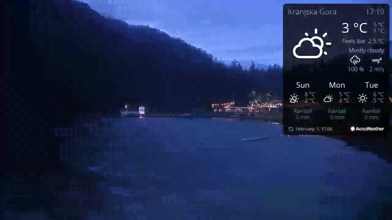 A mostly cloudy nighttime scene shows a dark body of water reflecting the lights of buildings and trees on a hill under a twilight sky.