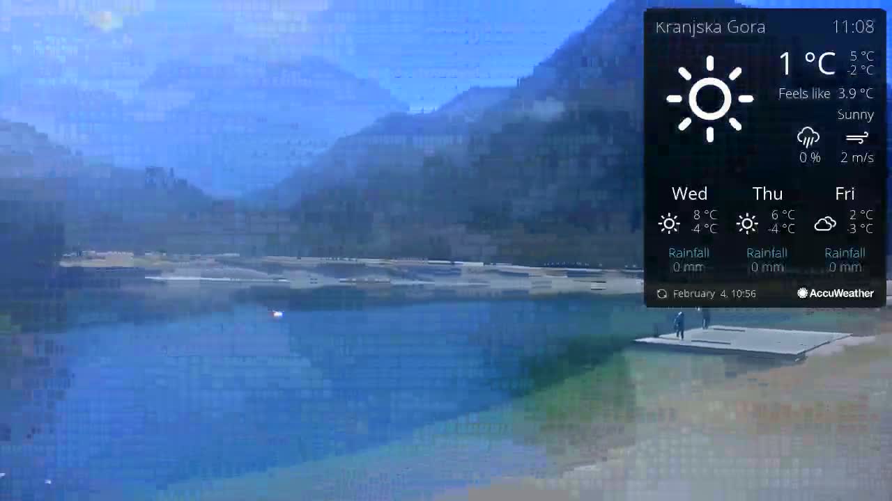 A sunny, calm day is shown overlooking a body of blue water next to a flat, gray dock with a few people standing on it, and mountains in the background.
