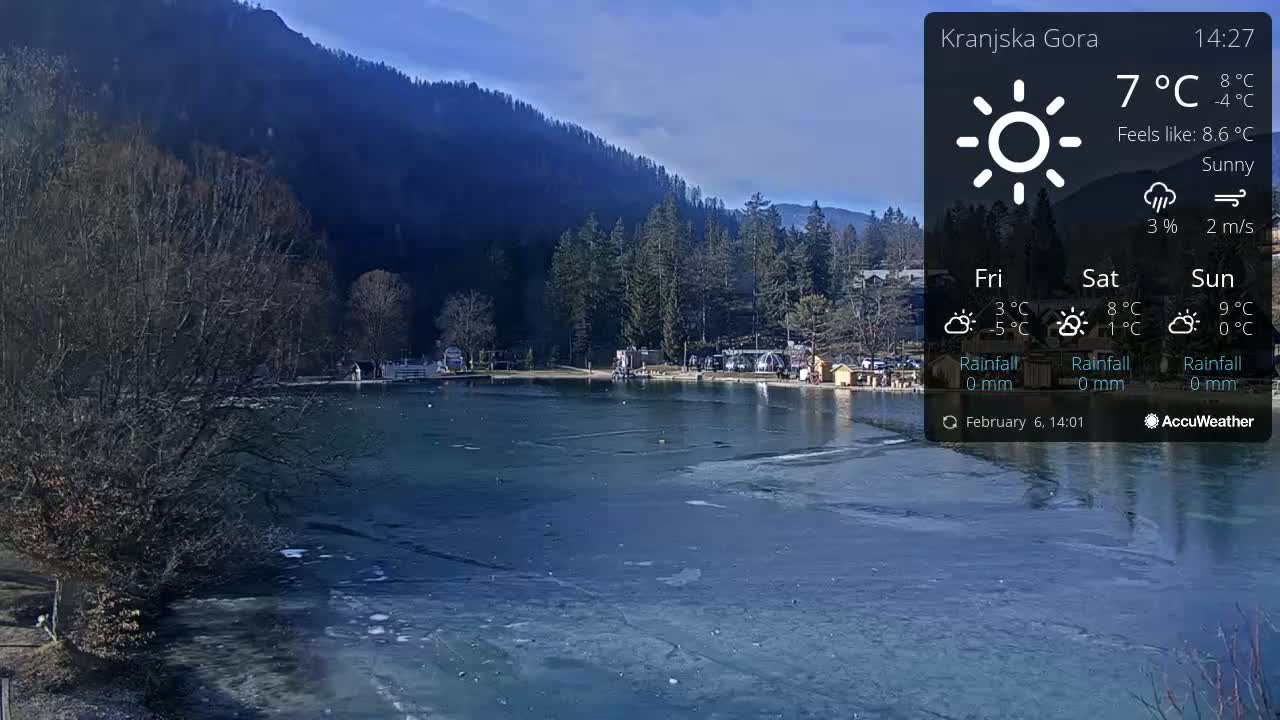 A partially frozen lake is situated in front of a mountainous forest under sunny skies.