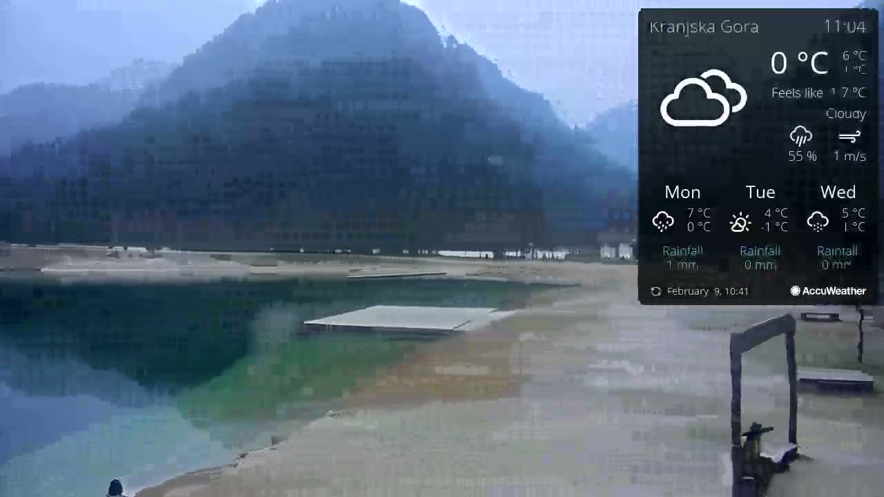 A cloudy, calm day shows a body of dark green water near a shore with a floating dock and mountains in the background.