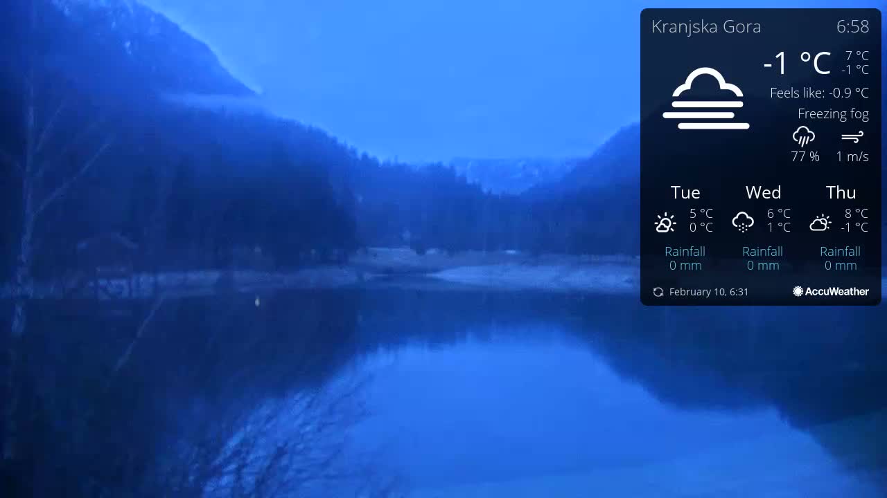 A calm lake reflects a dark mountain range under a freezing fog in the pre-dawn hours.