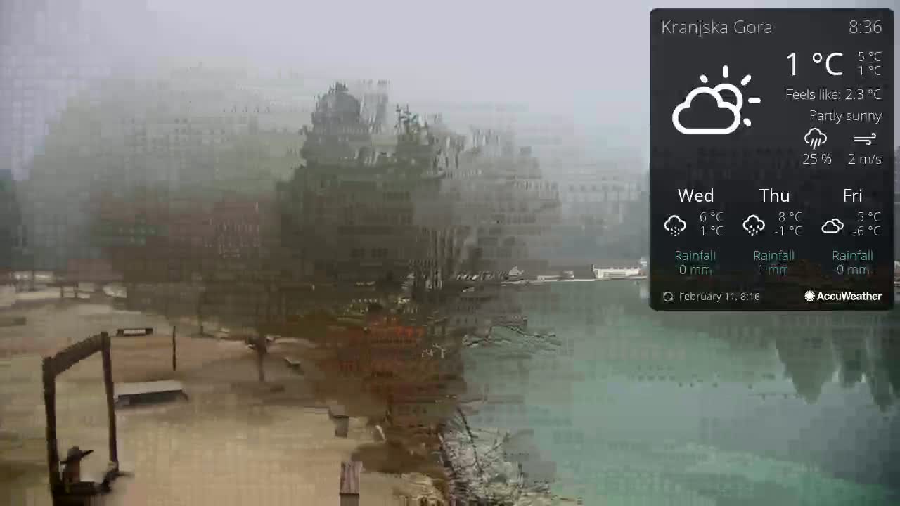 A foggy, partly sunny scene shows a body of water reflecting a hazy, indistinct town or city in the background, with a park-like area with benches and a wooden structure in the foreground.