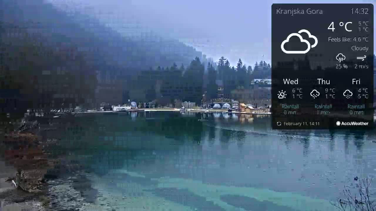 A calm lake reflects a cloudy sky and a town nestled at the base of hazy, snow-dusted mountains.