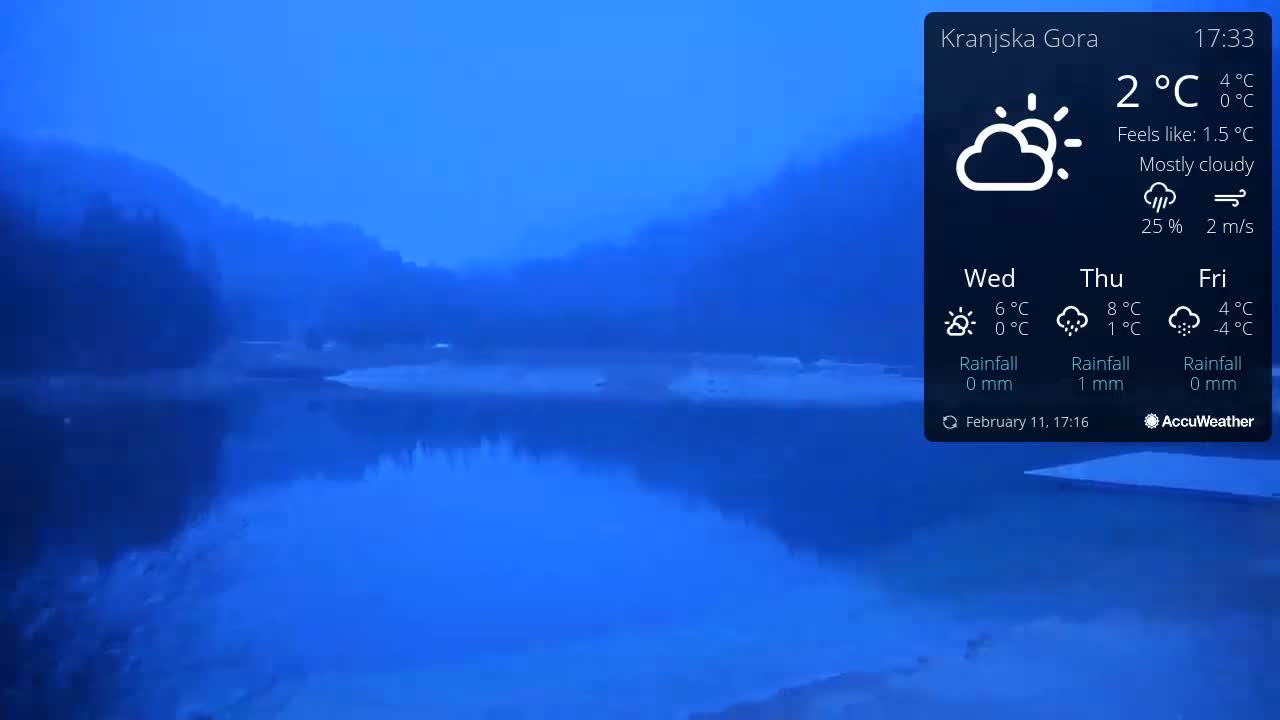 A mostly cloudy, twilight scene shows a calm body of water reflecting a hazy, dark blue sky and hills in the background.