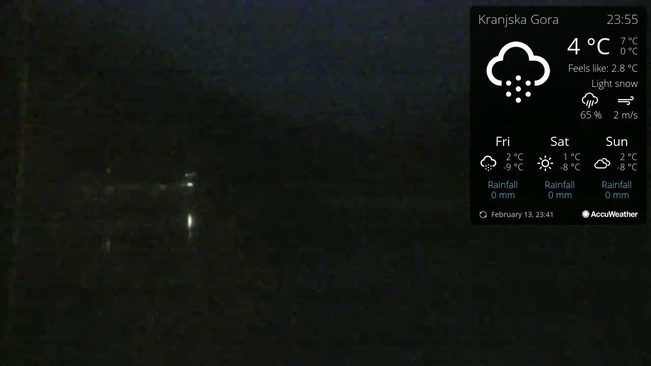 A dark, nighttime outdoor scene with light snow falling, showing a dimly lit area with faint lights in the distance.
