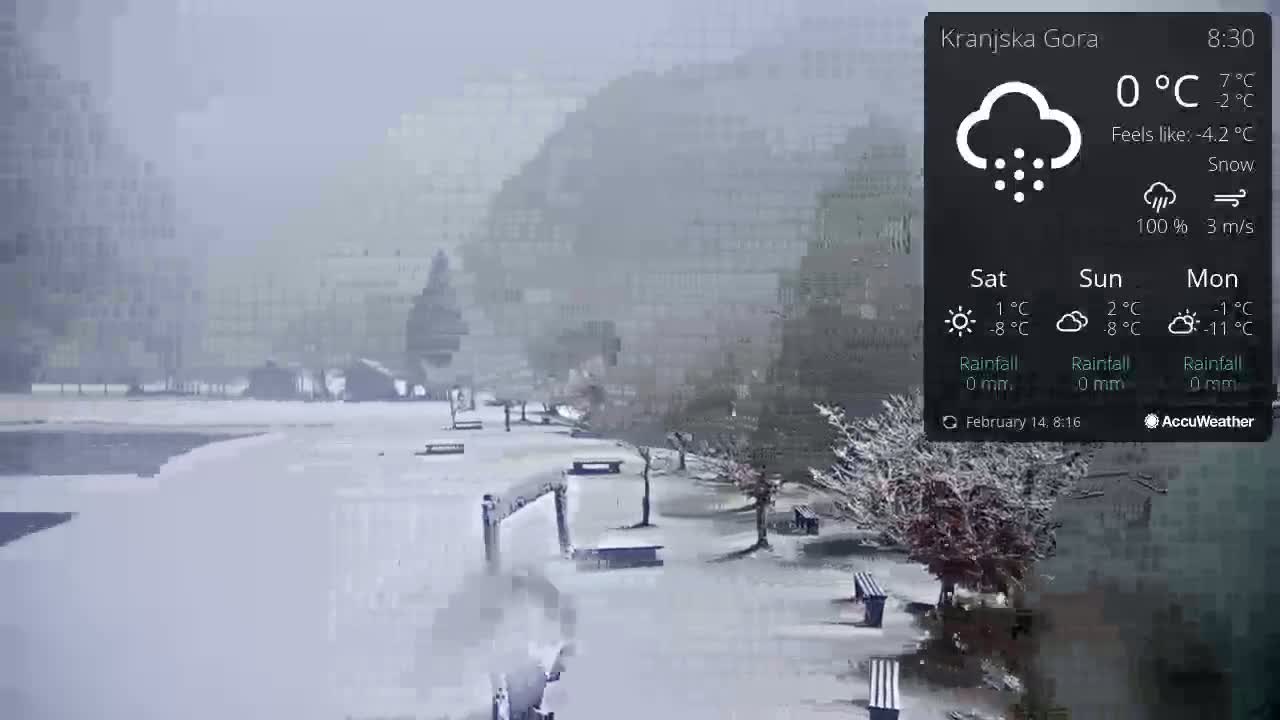 A snow-covered park with benches and trees is seen on a snowy day, with buildings in the background.