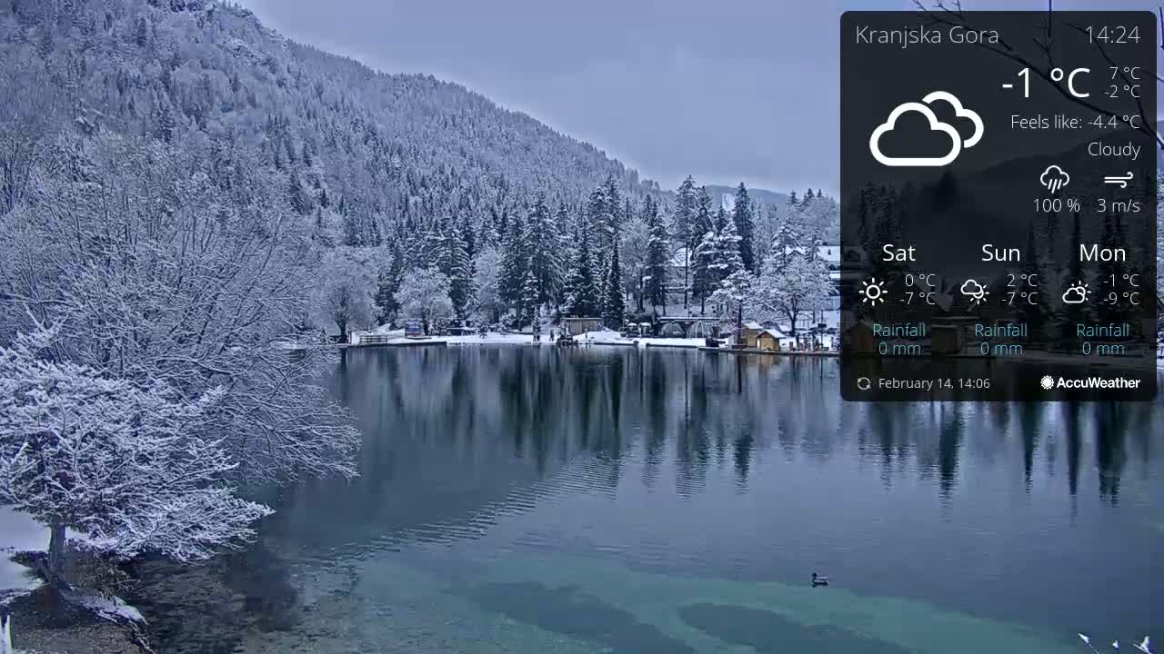 A snow-covered lake reflects a snow-dusted mountainside and small village under a cloudy sky.