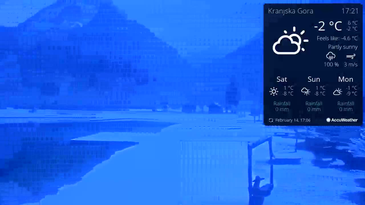 A partly sunny, cold scene with a body of water reflecting a snow-covered landscape and buildings under a blue sky.