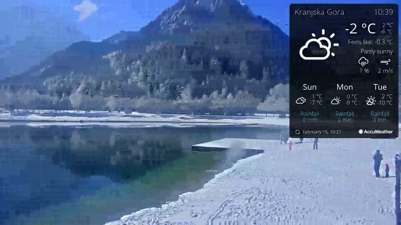 A partly sunny, cold day shows a snow-covered shoreline beside a calm lake, with a mountain in the background.