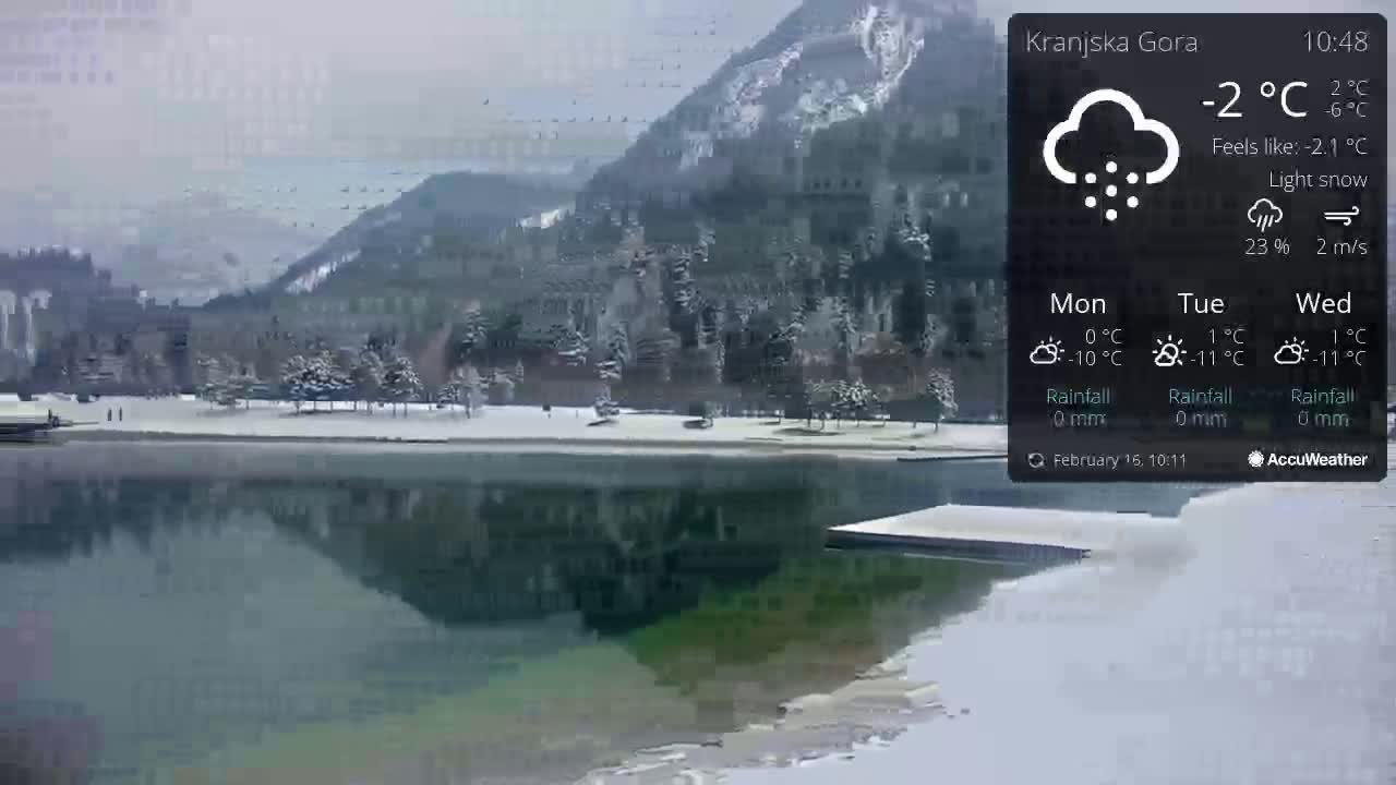 A snow-covered landscape with a calm lake reflecting a snow-dusted town and mountains under a light snow.