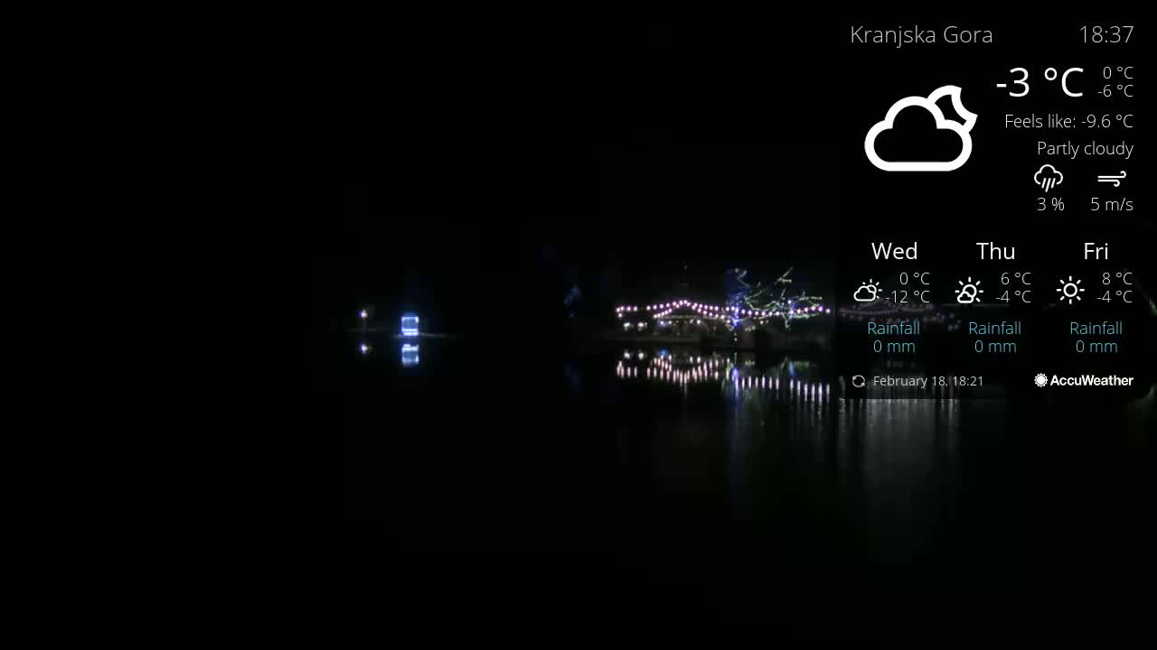 A nighttime view of a calm body of water reflecting lights from a distant, festively lit area under a partly cloudy sky.