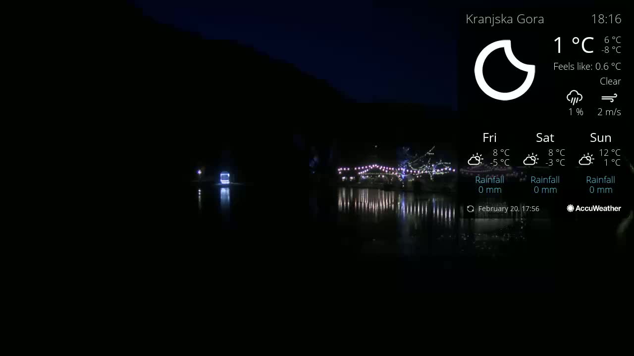 A nighttime scene shows a body of water reflecting lights from a structure on the opposite shore under clear skies.