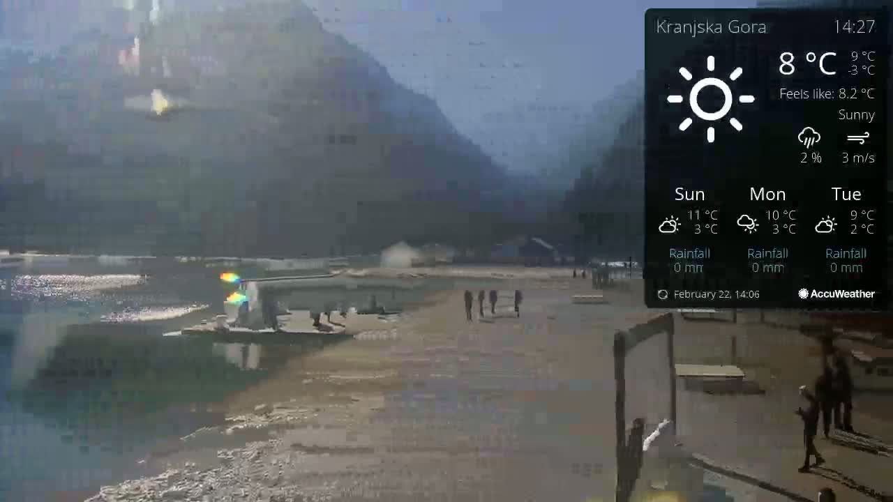 A sunny beach scene with a calm body of water, mountains in the background, and several people walking along the shore.