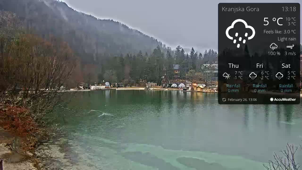 A calm, green lake sits before a misty, forested mountain range under a light rain.