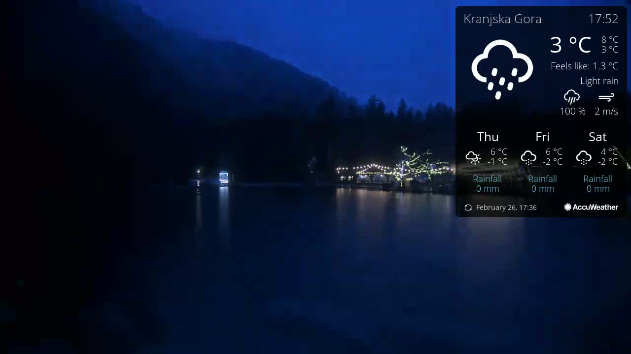 A dark, calm lake reflects the lights of a structure decorated with string lights, under a night sky with light rain.