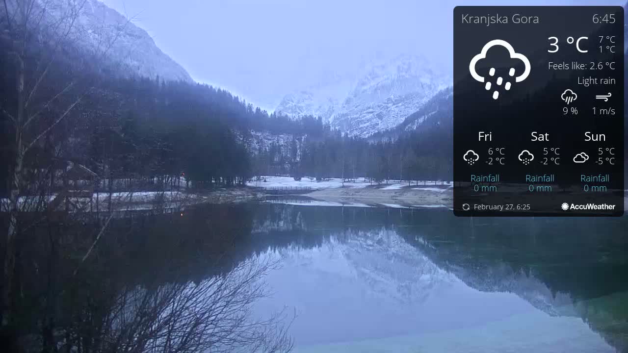 A calm lake reflects snow-capped mountains and a forest under a light rain.