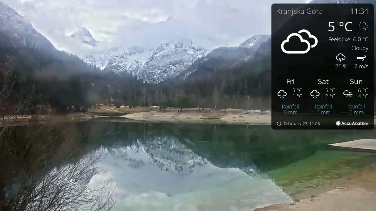 A calm lake reflects snow-capped mountains under a cloudy sky.