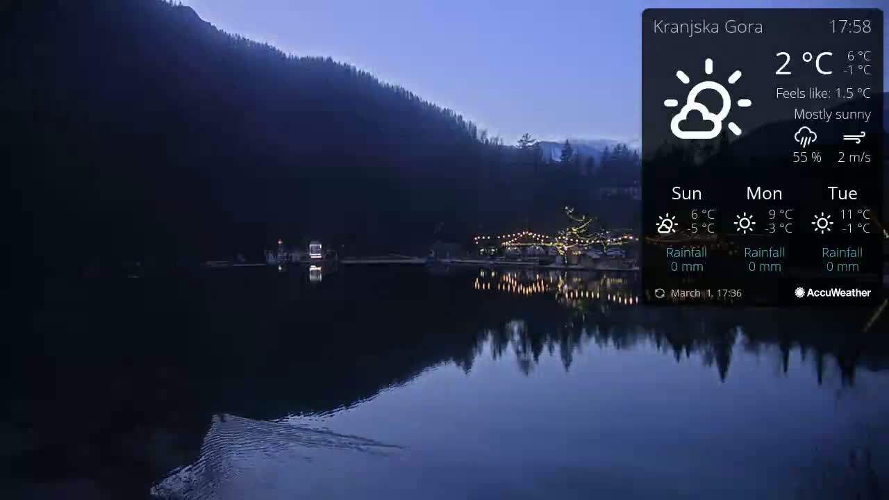 A mostly sunny twilight scene shows a calm lake reflecting a dark mountainside and illuminated shoreline buildings.