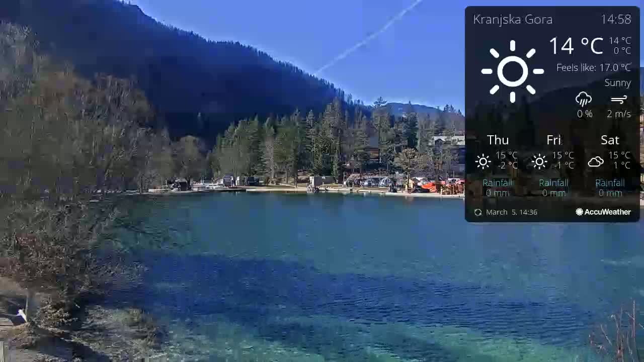 A sunny day shows a calm, clear blue lake nestled in a valley surrounded by dark green mountains.