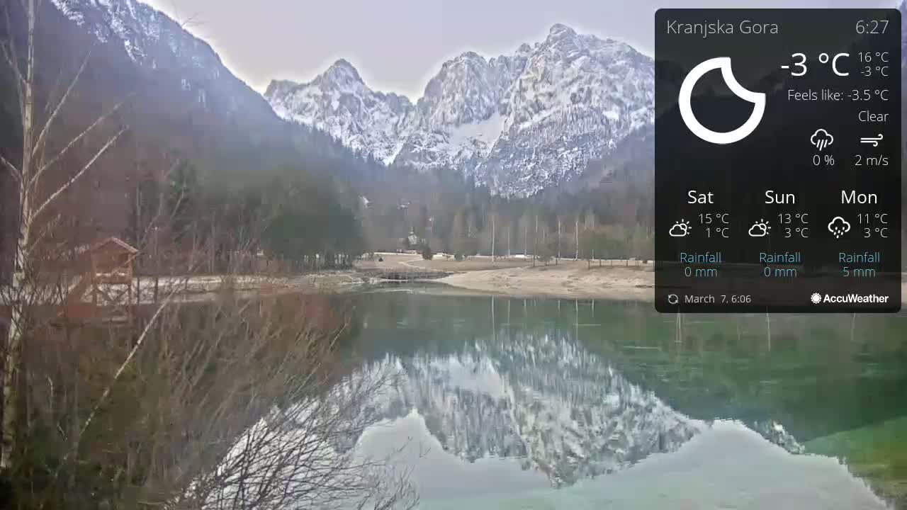 A calm, clear lake reflects snow-capped mountains under a gray sky.