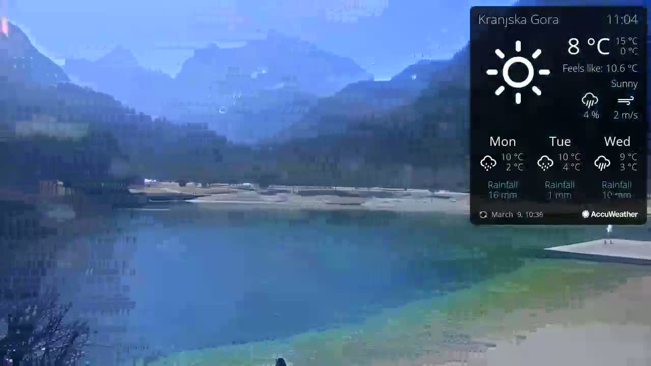 A sunny, calm day is shown overlooking a tranquil lake surrounded by mountains.