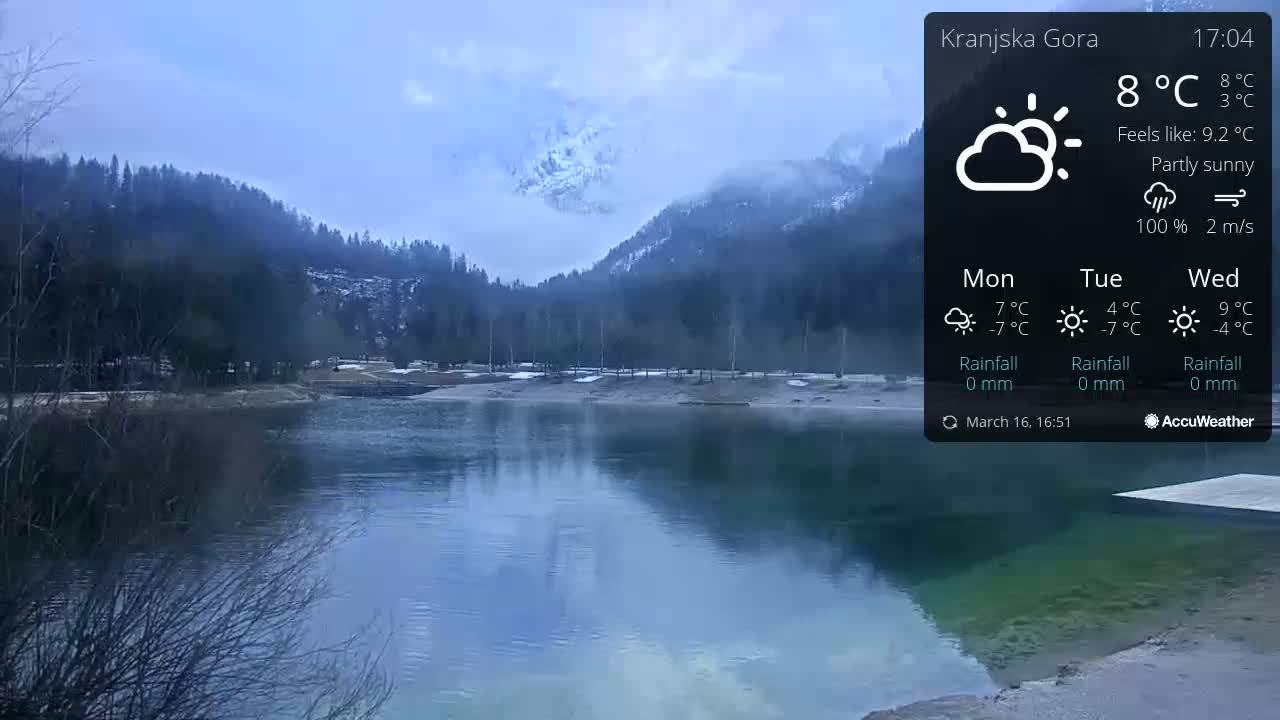 A partly sunny day reveals a calm lake reflecting mountains shrouded in mist, with a shoreline showing patches of snow.