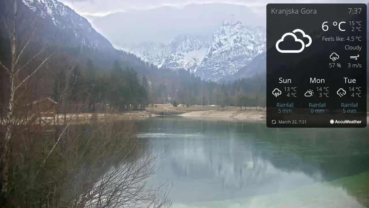 A calm lake reflects snow-capped mountains under a cloudy sky.