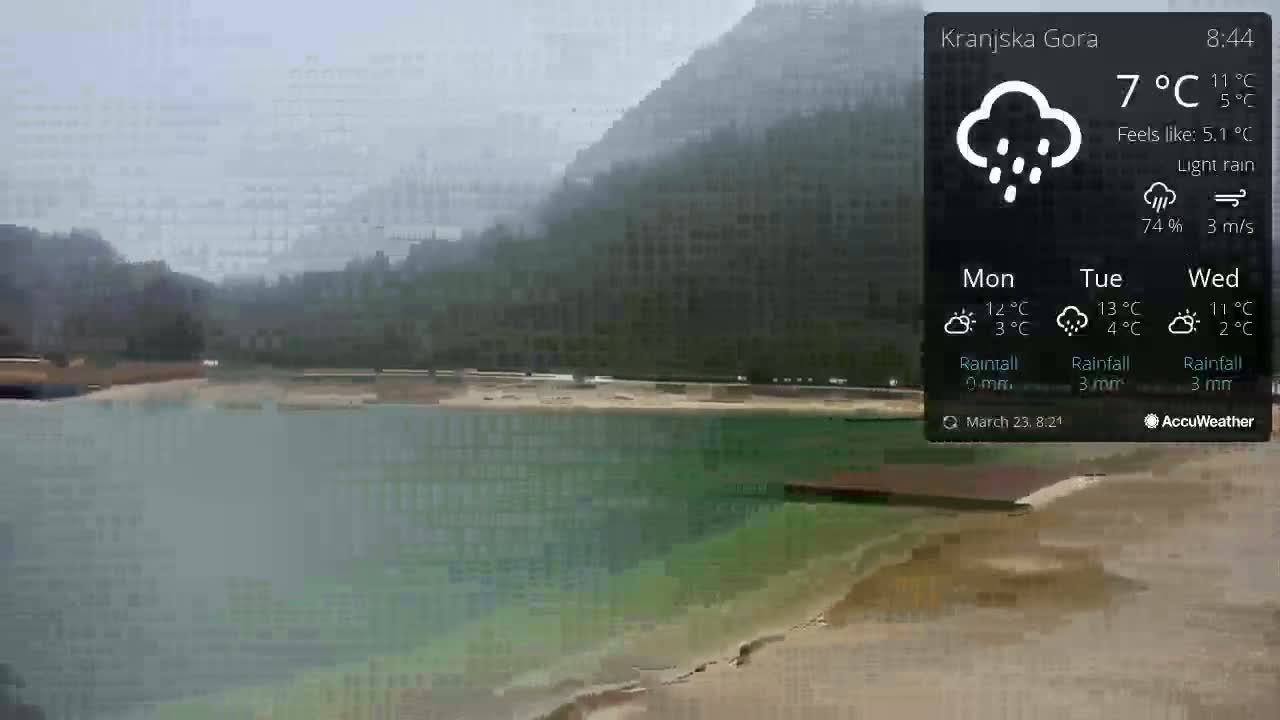 A lake with a sandy shore is visible under a light rain, with mountains in the background.