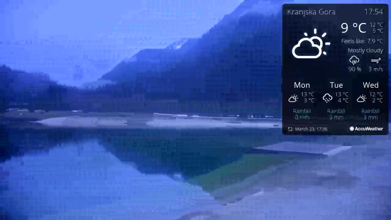 A mostly cloudy, cool day shows a calm lake reflecting mountains under a blue sky.