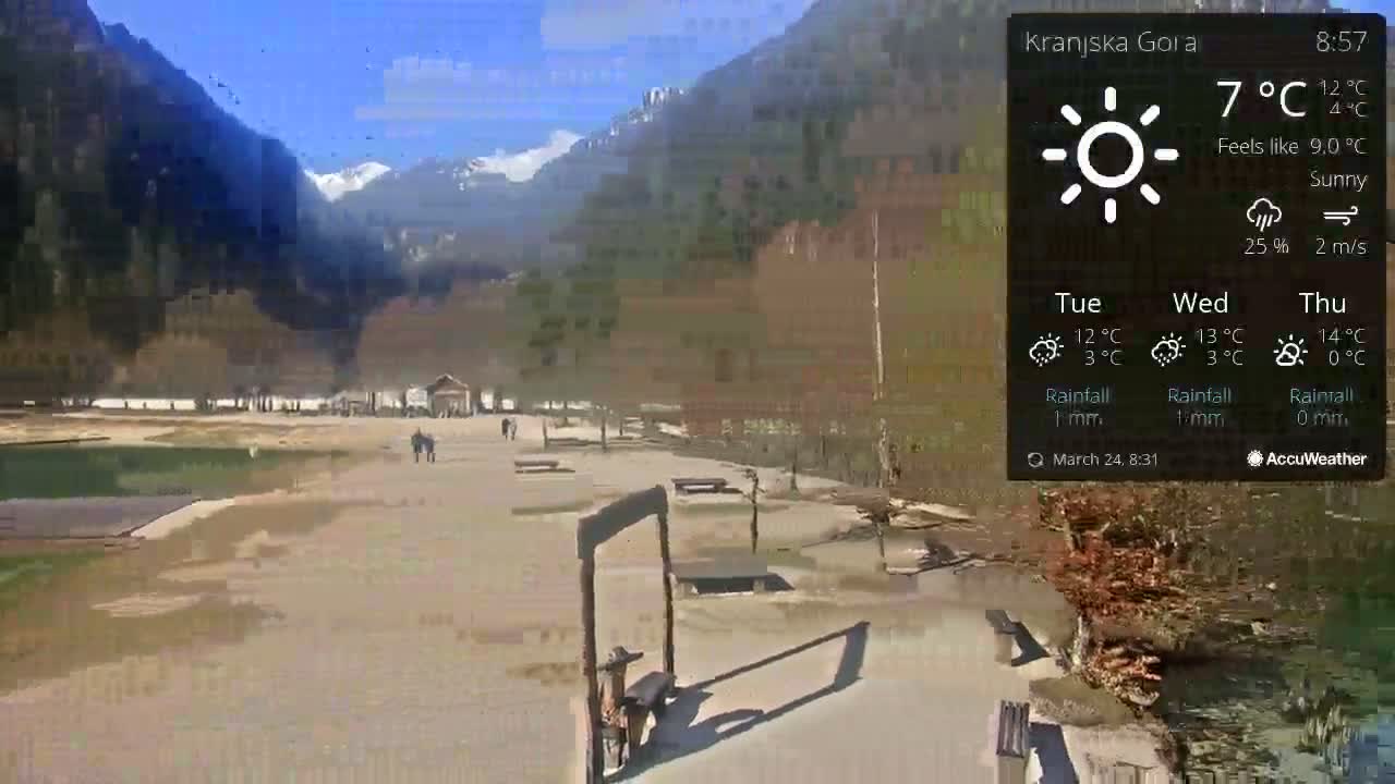 A sunny outdoor scene shows a paved area with picnic tables and a few people walking, near a calm body of water and mountains in the background.