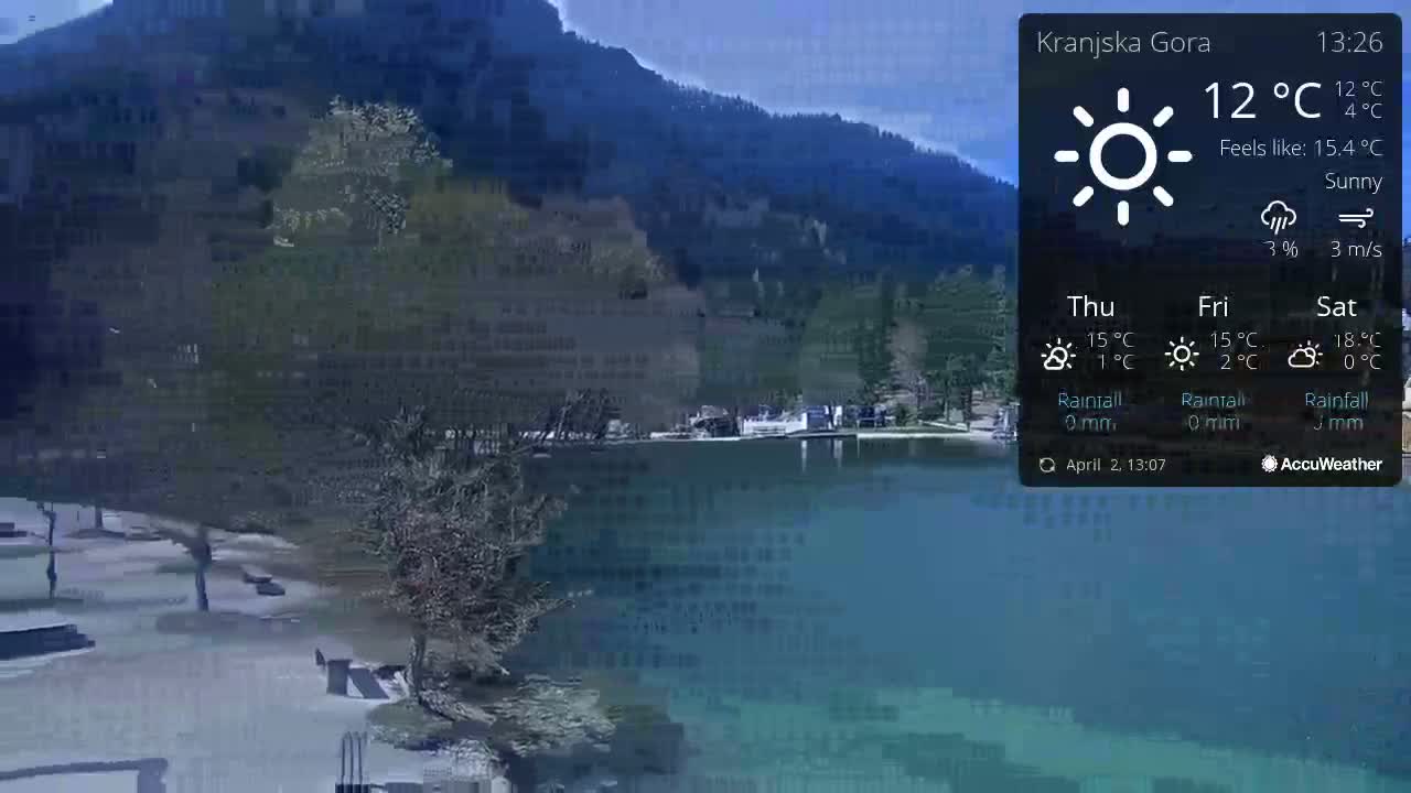 A sunny view of a lake and mountains with some trees and buildings along the shore.
