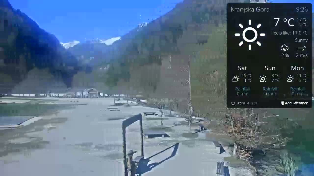 A sunny outdoor scene shows a flat, paved area with a few park benches and trees near a body of water, all set against a backdrop of mountains with some snow visible on their peaks.