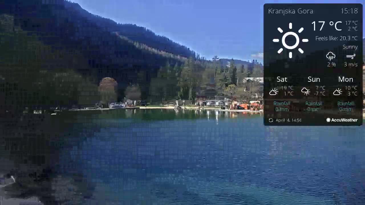 A calm, sunny lake reflects a mountainside covered in dark green trees.