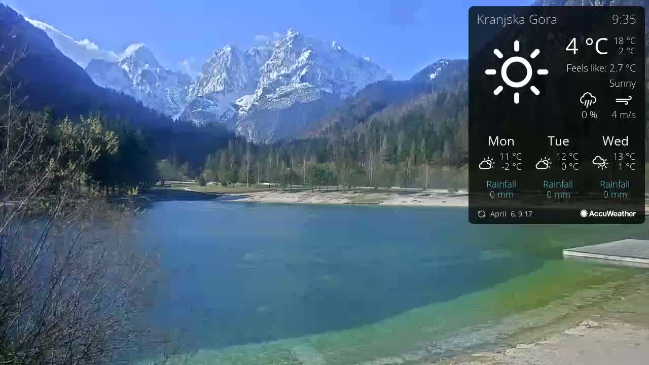A tranquil lake with clear, teal-green water sits before a backdrop of snow-capped mountains under a sunny sky.