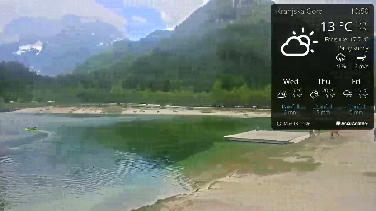 A partly sunny view of a lake with a sandy shore, surrounded by green mountains, and a small floating dock.