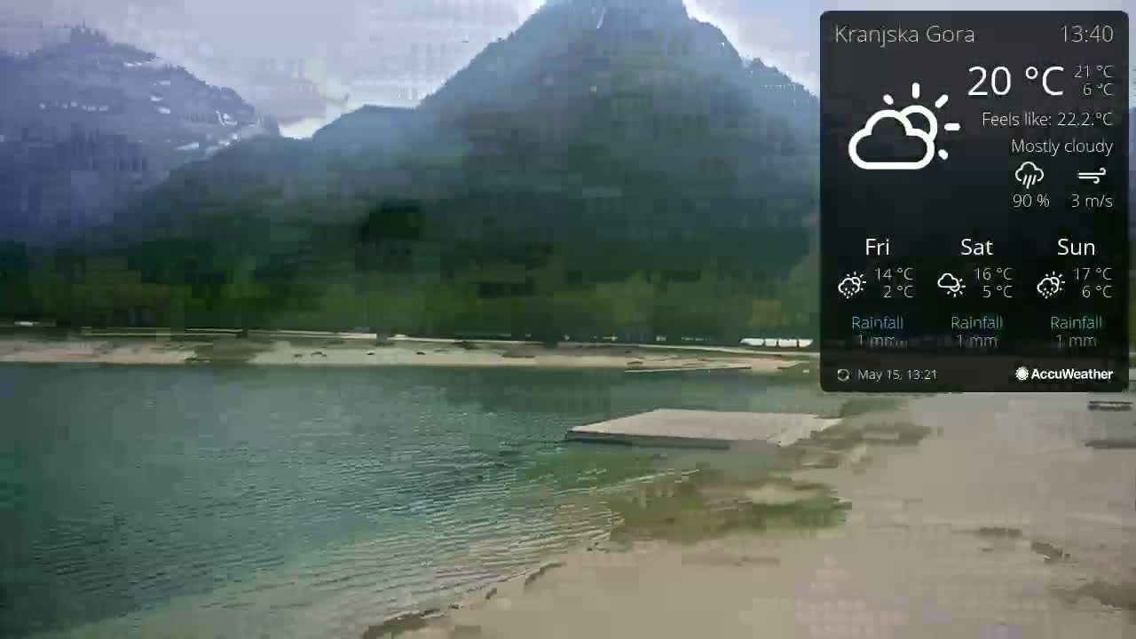 A mostly cloudy day shows a calm lake with a small dock in front of a mountainous, green shoreline.